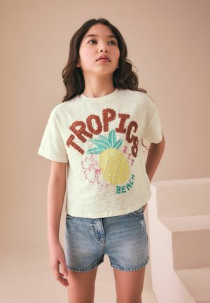 Ragazza con capelli lunghi e scuri che indossa una maglietta bianca con la scritta "TROPICS BEACH" e un disegno di ananas e pantaloncini di jeans, in piedi all'interno vicino alle scale.