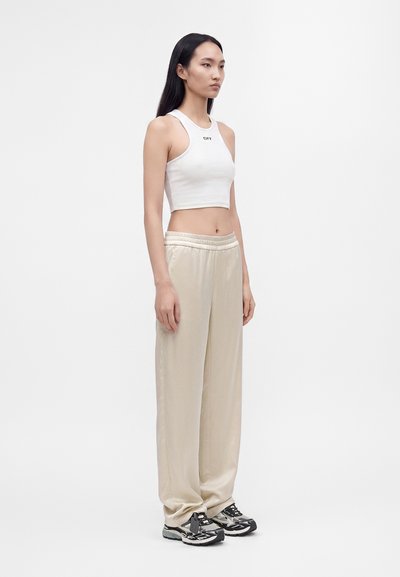 Femme aux longs cheveux noirs portant un débardeur court blanc, un pantalon ample beige et des baskets noires et blanches, debout devant un fond uni.