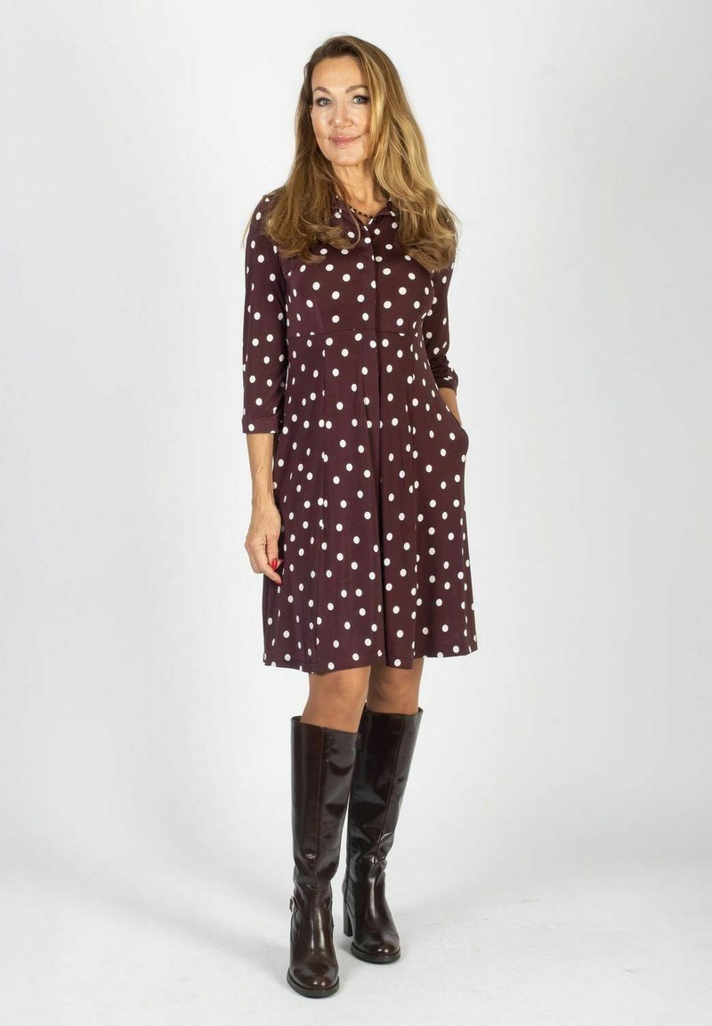 Brązowa sukienka do kolan w białe kropki, z przodem na guziki i długimi rękawami. Posiada boczne kieszenie oraz lekko rozkloszowany dół.