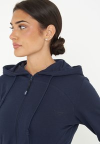 Felpa con cerniera blu navy, realizzata in un tessuto morbido, con cordino regolabile e dettaglio del logo sul petto. Presenta un cappuccio foderato e una vestibilità aderente.