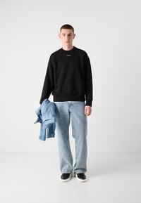 Sudadera negra con logo, jeans de mezclilla azul claro, sosteniendo una chaqueta de mezclilla. Zapatos negros sin cordones, textura suave, ajuste relajado en general.