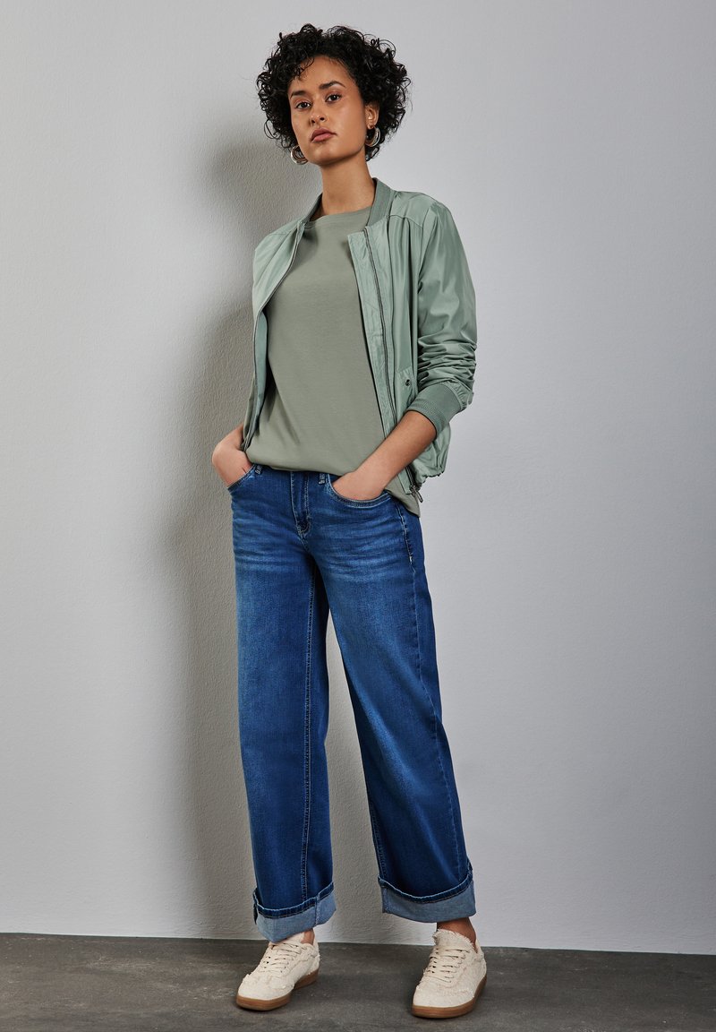 Giacca bomber verde chiaro, t-shirt oliva aderente, jeans blu a gamba larga con risvolti e sneakers beige. Design semplice e vestibilità rilassata.