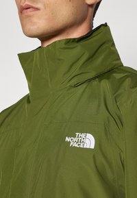 Grønn vanntett jakke med høy krage, med en hvit logo på brystet og sømdetaljer. Glatt tekstur, stilrent design.
