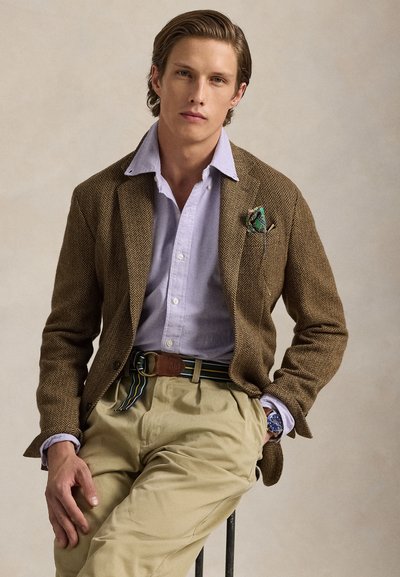 Młody mężczyzna z zaczesanymi do tyłu włosami, ubrany w brązową marynarkę w jodełkę, jasnofioletową koszulę, beżowe spodnie i paskiem w paski, siedzi na stołku.