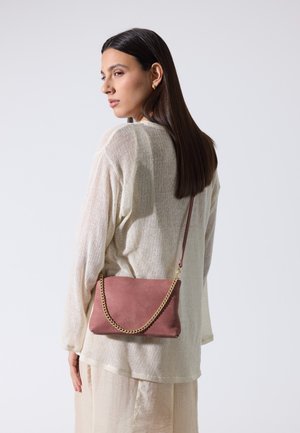 Femme aux longs cheveux foncés portant un pull crème et une jupe, portant un sac à bandoulière mauve avec une chaîne dorée, face à gauche.