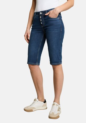 Frau trägt dunkelblaue Denim-Knielange Shorts mit Knopfleiste, weiße Sneaker und ein weißes ärmelloses Top, steht mit einer Hand in der Tasche.