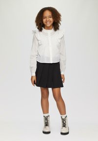 Blouse blanche avec un col à volants, un devant boutonné et des détails en petites plis, assortie d'une jupe plissée noire et de bottines beiges à lacets.