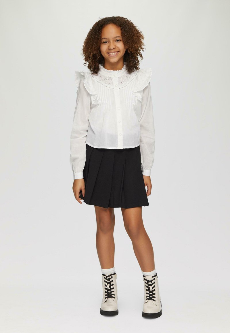 Blouse blanche avec un col à volants, un devant boutonné et des détails en petites plis, assortie d'une jupe plissée noire et de bottines beiges à lacets.