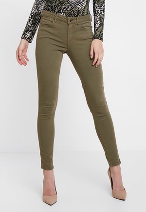 Kvinde iført olivengrønne tætsiddende bukser og beige højhælede sko, kombineret med en langærmet top med slangeskindsmønster.