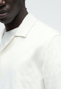 Gros plan sur une personne portant une chemise en tissu texturé blanc avec un col et un sous-vêtement blanc, sur un fond uni.