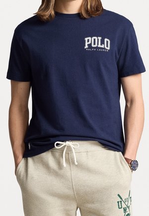 Homme portant un t-shirt Polo Ralph Lauren bleu marine et un pantalon de survêtement beige clair avec cordon de serrage, mains dans les poches, et montre au poignet gauche.