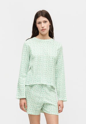 Jonge vrouw met lichtgroene top met bloemenprint en lange mouwen en bijpassende shorts, staand tegen een effen witte achtergrond.