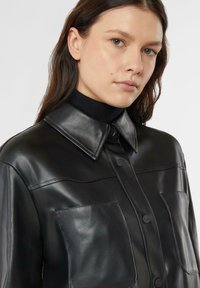 Czarna skórzana koszula-kurtka z zapinanym na guziki frontem, dużymi kieszeniami na piersi i spiczastym kołnierzem. Gładka faktura i dopasowany krój.