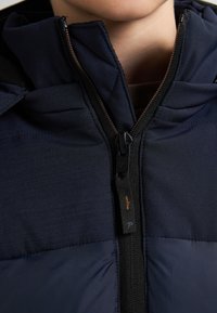 Jachetă bleu marin cu guler înalt, material texturat, secțiuni contrastante mate și lucioase, și un fermoar frontal cu un tab negru.