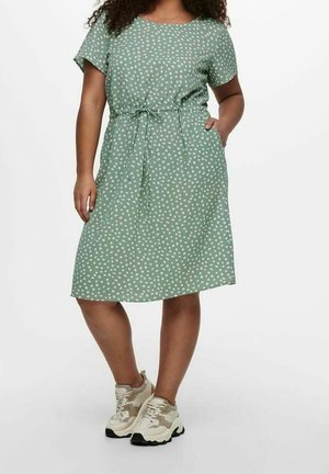 Frau trägt ein grünes knielanges Kleid mit Punkten und kurzen Ärmeln sowie beige klobige Sneakers, steht mit einer Hand in der Tasche.