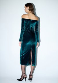 Turkusowa welurowa sukienka na ramiączkach z długimi rękawami, z rozcięciem z tyłu i dopasowanym krojem. Czarne szpilki z paskami na kostkę dopełniają stylizację.