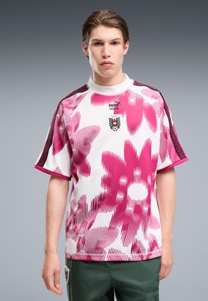 Tânăr purtând un tricou de fotbal Puma alb și roz cu model și logo OFB, stând pe un fundal gri simplu.