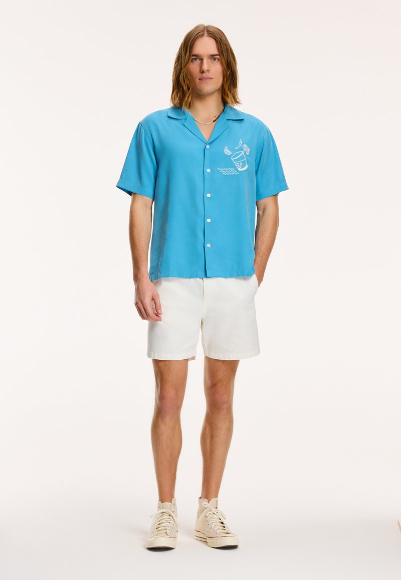 Lyseblå kortærmet skjorte med krave, der har et hvidt grafisk design på venstre side, parret med hvide shorts og beige sneakers.