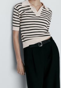 Pull polo à rayures crème et noires à manches courtes, texture côtelée, col en V, porté avec un pantalon noir et une ceinture noire.