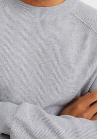 Jasnoszary sweter dzianinowy o prążkowanej fakturze, z okrągłym dekoltem i długimi rękawami. Materiał wydaje się miękki i elastyczny, z widocznymi detalami szwów.