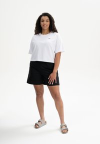 Weißes, kurzes T-Shirt mit kleinem Logo, schwarze Shorts und graue Sandalen. Der Stoff ist weich, und das Design ist einfach und lässig.