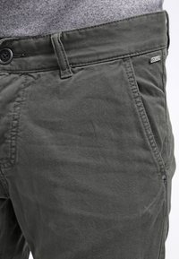 Zelené bavlněné chinos kalhoty mají hladkou strukturu, zapínání na knoflíky, přední kapsy a jemné značkování na pásku.