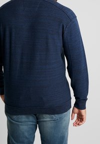 Navy blue textured sweater with long sleeves, ribbed cuffs, and hem. Made from knit material, featuring subtle horizontal patterns.