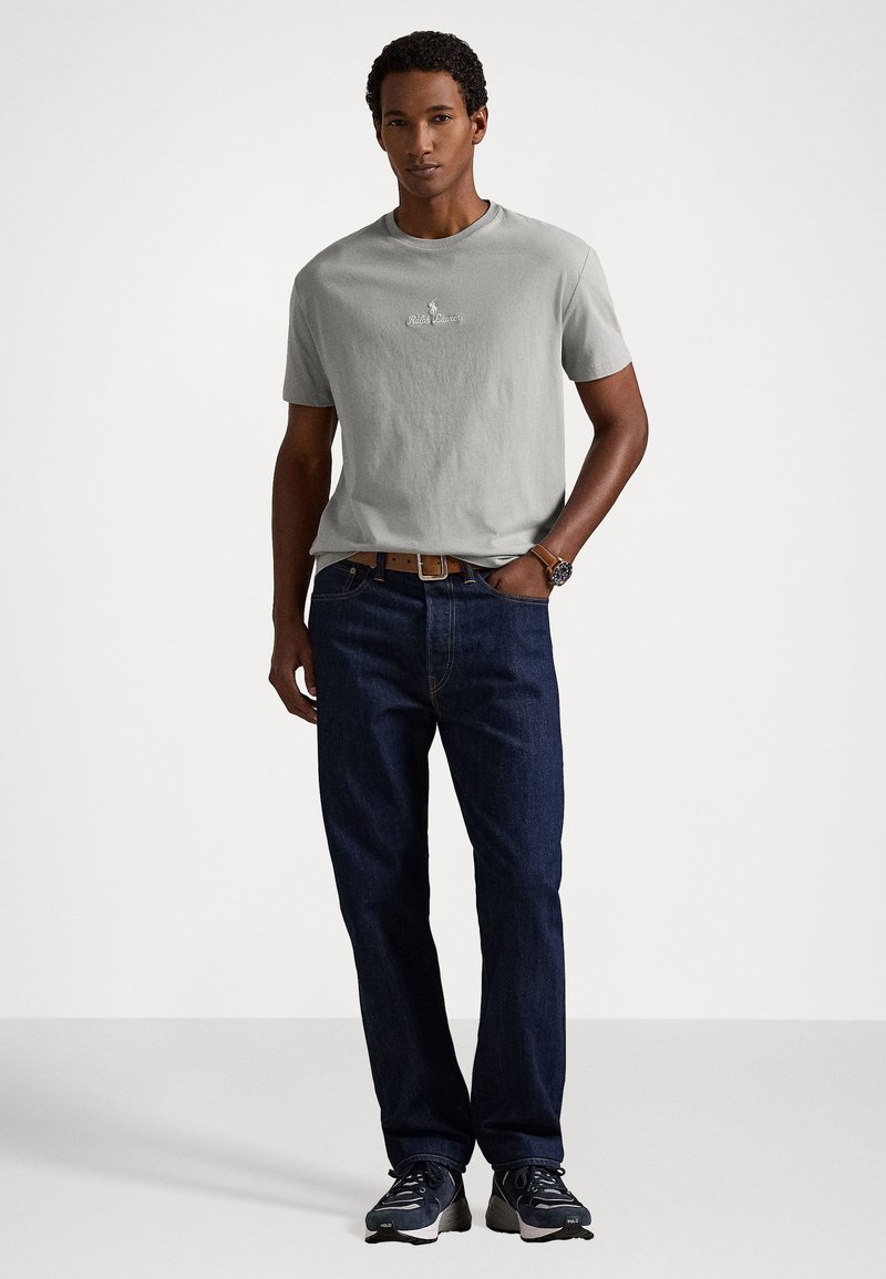 T-shirt in cotone grigio chiaro con scollatura a girocollo e logo sul petto, abbinato a jeans blu scuro e scarpe da ginnastica nere. Il modello è in posizione frontale.