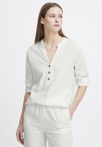 Blouse en lin blanc avec un décolleté en V et trois boutons décoratifs. Manches retroussables et coupe décontractée. Texture douce avec une apparence légère.