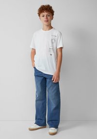 Camiseta blanca de manga corta con estampados gráficos, combinada con pantalones vaqueros anchos azules y zapatillas blancas con suela beige.