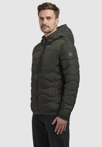 Dunkelgrüne gepolsterte Jacke mit Kapuze, ausgestattet mit einem Reißverschluss, seitlichen Taschen und einem Wellenmuster auf der Oberfläche. Strukturstoff.