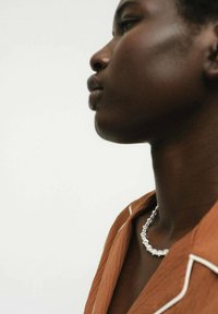 Collana in argento indossata attorno al collo, con una finitura testurizzata. Lo sfondo è neutro, mettendo in risalto l'accessorio e il colore ruggine della camicia.