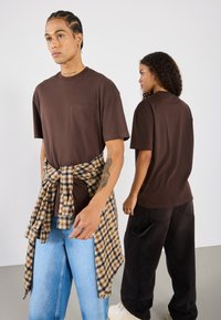 Camisetas marrones de gran tamaño con un bolsillo en el pecho, combinadas con jeans azul claro y una camisa de franela a cuadros atada a la cintura. Pantalones negros por detrás.