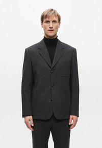 Blazer grigio in tessuto strutturato, con colletto a punta, due tasche anteriori e tre bottoni, abbinato a un dolcevita nero.