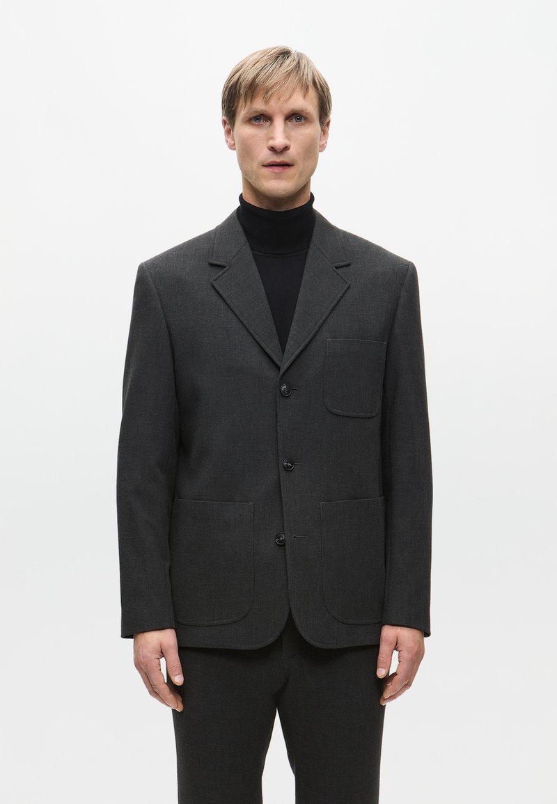 Blazer grigio in tessuto strutturato, con colletto a punta, due tasche anteriori e tre bottoni, abbinato a un dolcevita nero.