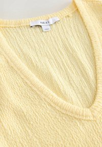 Tissu jaune doux et texturé avec un col en V et une étiquette blanche portant les inscriptions "NEXT" et "Fabriqué en Turquie".