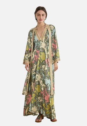 Mujer que lleva un vestido largo y suelto y un abrigo abierto a juego con patrones de paisley multicolor en verde pastel, rosa, azul y crema.