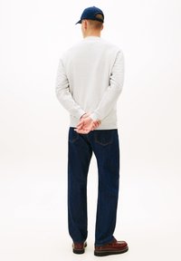 Ανοιχτό γκρι φούτερ, σκούρο μπλε τζιν, καφέ παπούτσια, στέκεται με τα χέρια πίσω από την πλάτη, απεικονίζεται από πίσω πάνω σε απλό φόντο.