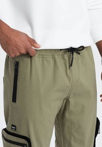 Pantalons cargo vert olive avec poches zippées noires et taille élastique. Présente des cordons de serrage et un patch logo sur la cuisse.
