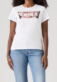 Žena oblečená v bílém tričku Levi's s růžovým květinovým logem a světlomodrých džínách, stojí s uvolněnýma rukama podél těla.