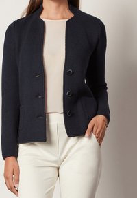 Mörkblå stickad kofta med rund halsringning, fyra framknappar, två sidfickor och strukturerad tyg. Parat med en krämfärgad topp och ljusa byxor.