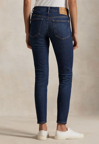 Persona che indossa jeans in denim slim-fit blu scuro e sneakers basse bianche, in piedi davanti a uno sfondo beige neutro.