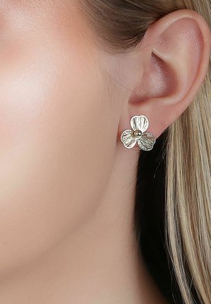 Goldener Ohrstecker in Form einer dreiblättrigen Blume, getragen am Ohrläppchen einer Person mit heller Haut und blondem Haar.
