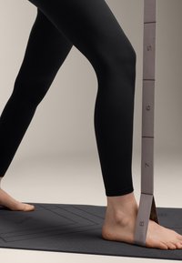 Fekete leggings, sima felülettel, részben láthatóak egy 5-től 8-ig terjedő számokkal megjelölt bézs szíj mellett. Egy láb pihen egy fekete jóga matracon.