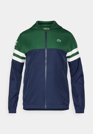 Giacca a zip verde e blu navy con cappuccio; materiale leggero, dettagli a strisce bianche sulle maniche e logo Lacoste sulla parte superiore sinistra.