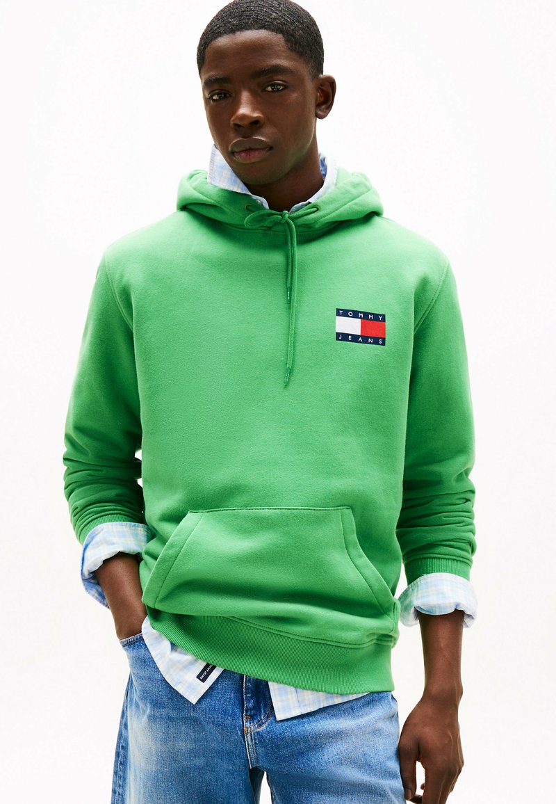 Grüne Baumwollkapuzenjacke mit einer Fronttasche und gerippten Bündchen, versehen mit einem Logo-Patch in Rot, Weiß und Blau. Darunter trägt er ein helles kariertes Hemd.