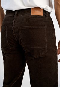 Brązowe spodnie z korduroju mają teksturowaną powierzchnię, pięć kieszeni oraz skórzaną metkę na pasie z wytłoczonym napisem "LINDBERGH".
