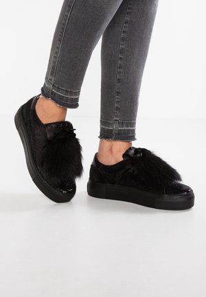 Czarne sneakersy na platformie z dużymi futrzanymi pomponami, noszone z szarymi obcieranymi obcisłymi jeansami na białej podłodze.