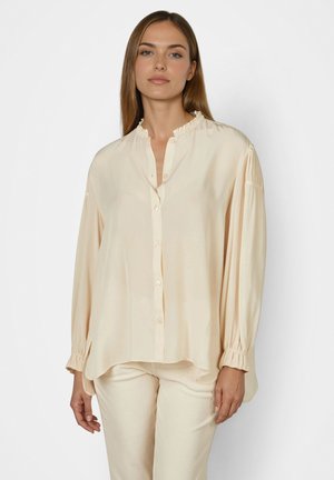 Woman wearing loose cream blouse with ruffled collar and cuffs, paired with matching cream pants, standing against a plain background.