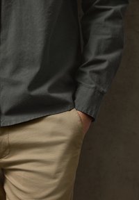 Rechte Hand in der Tasche einer beigefarbenen Hose, trägt ein dunkelgrünes, langärmliges Hemd mit strukturierter Stoffoberfläche vor einem dunklen neutralen Hintergrund.